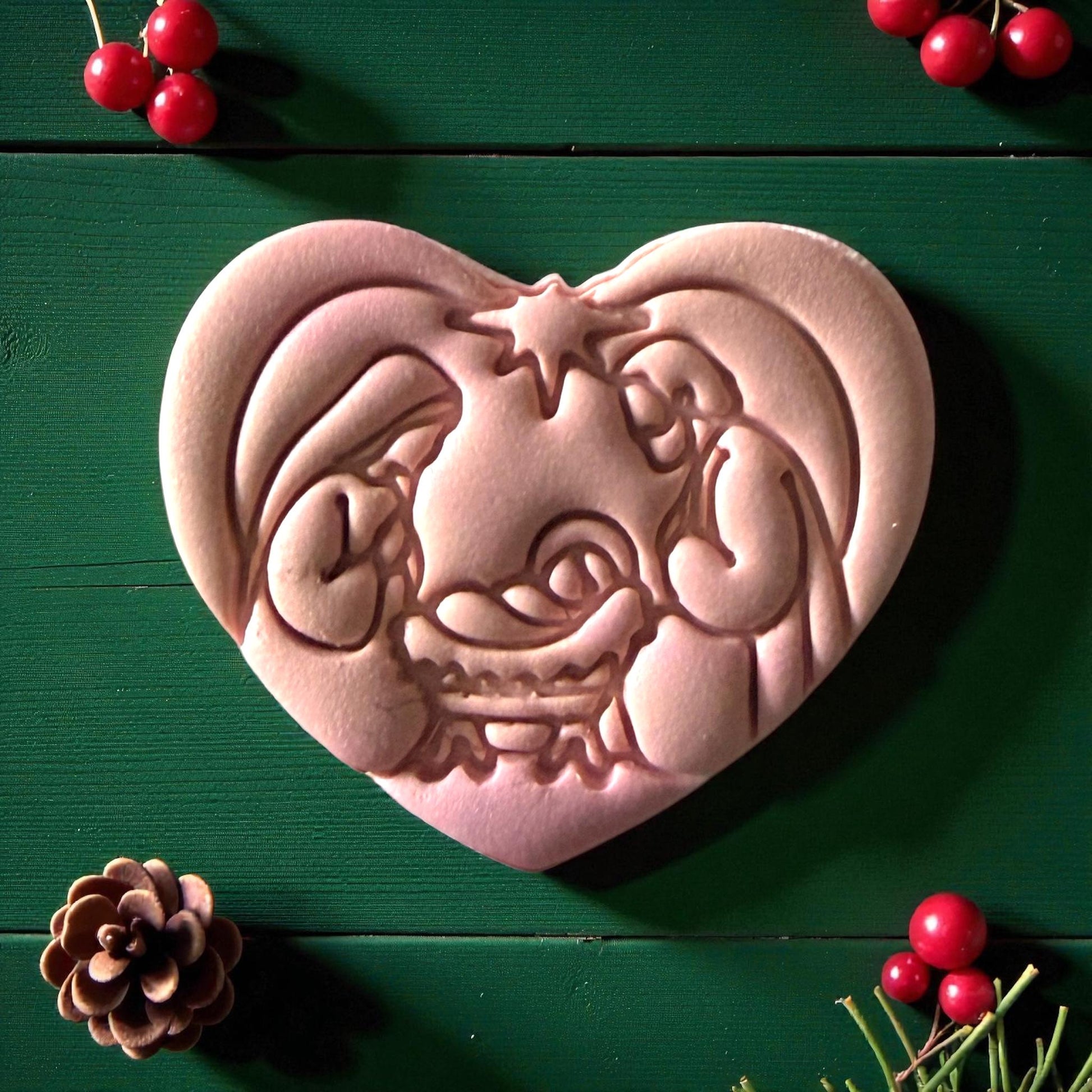 Close-up of pink nativity clay imprint shaped like a heart with pinecone and red berries nearby