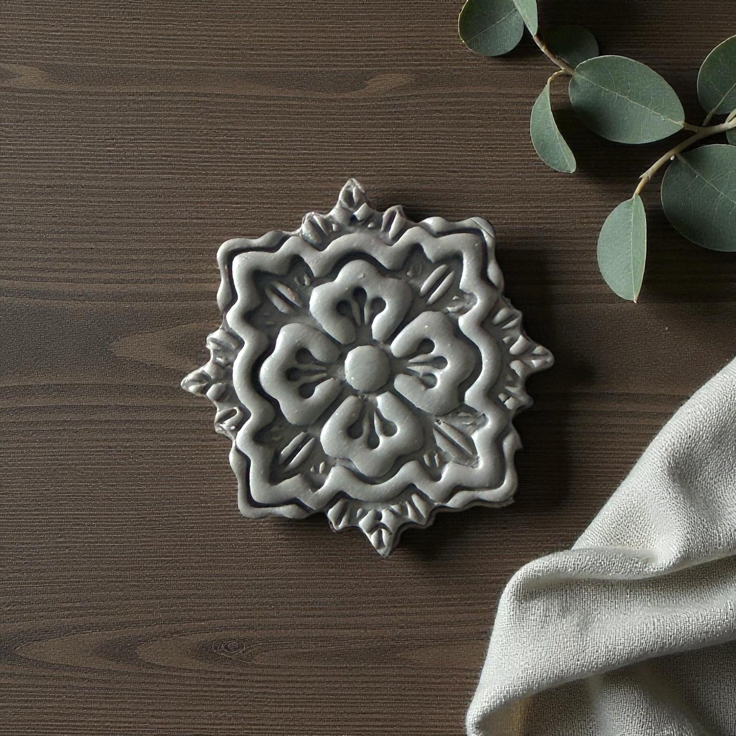 Gray floral medallion clay imprint on wood background with eucalyptus and beige fabric