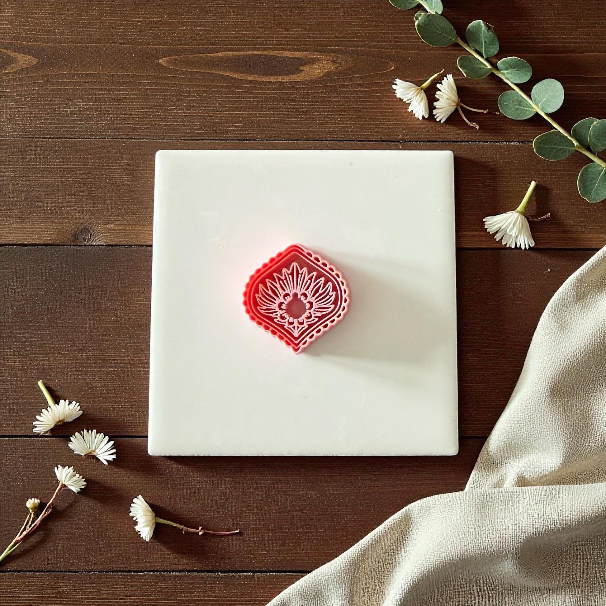 Pink diamond sunburst clay cutter beside imprint on white tile surrounded by greenery