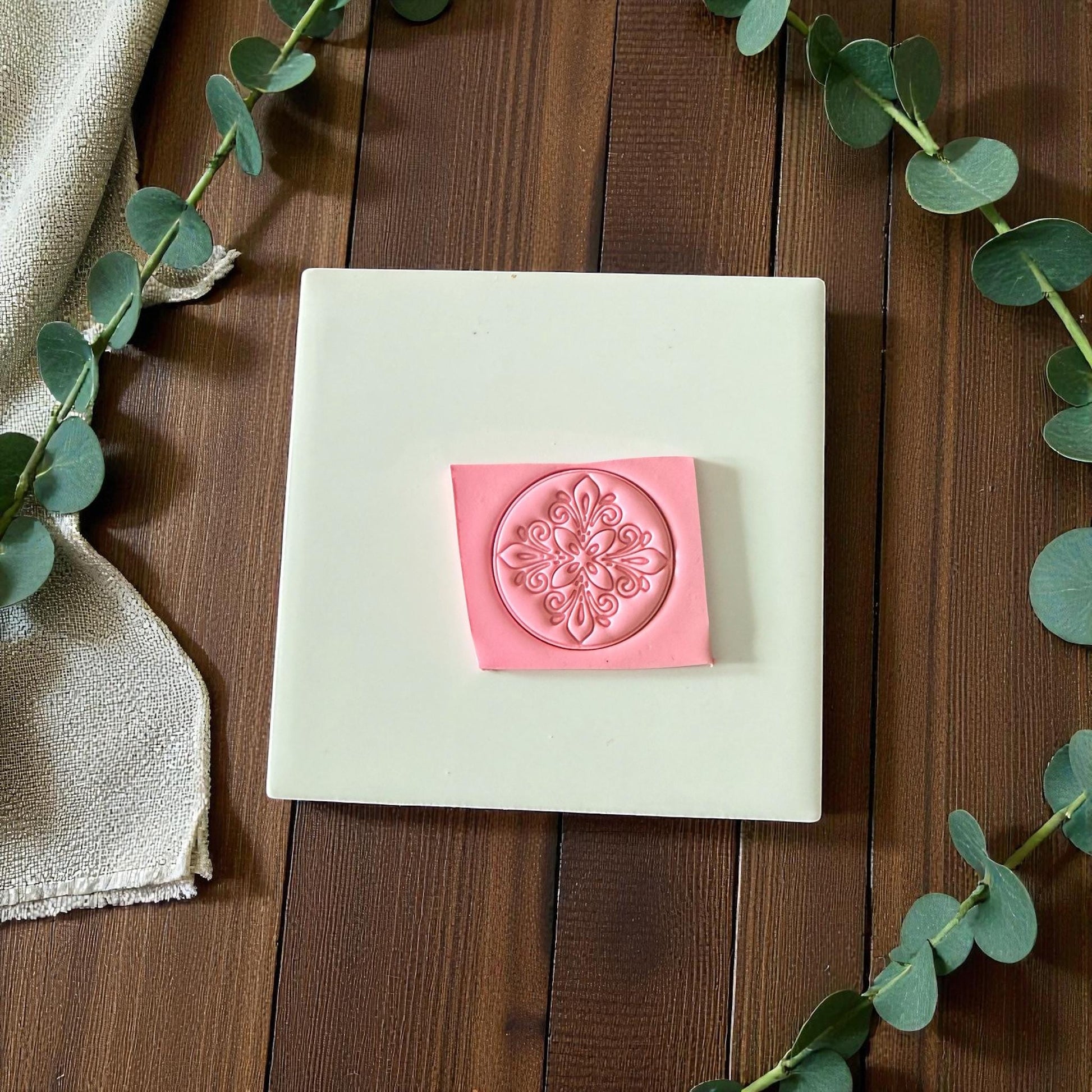 Round ornate clay imprint on square clay slab with eucalyptus and linen cloth