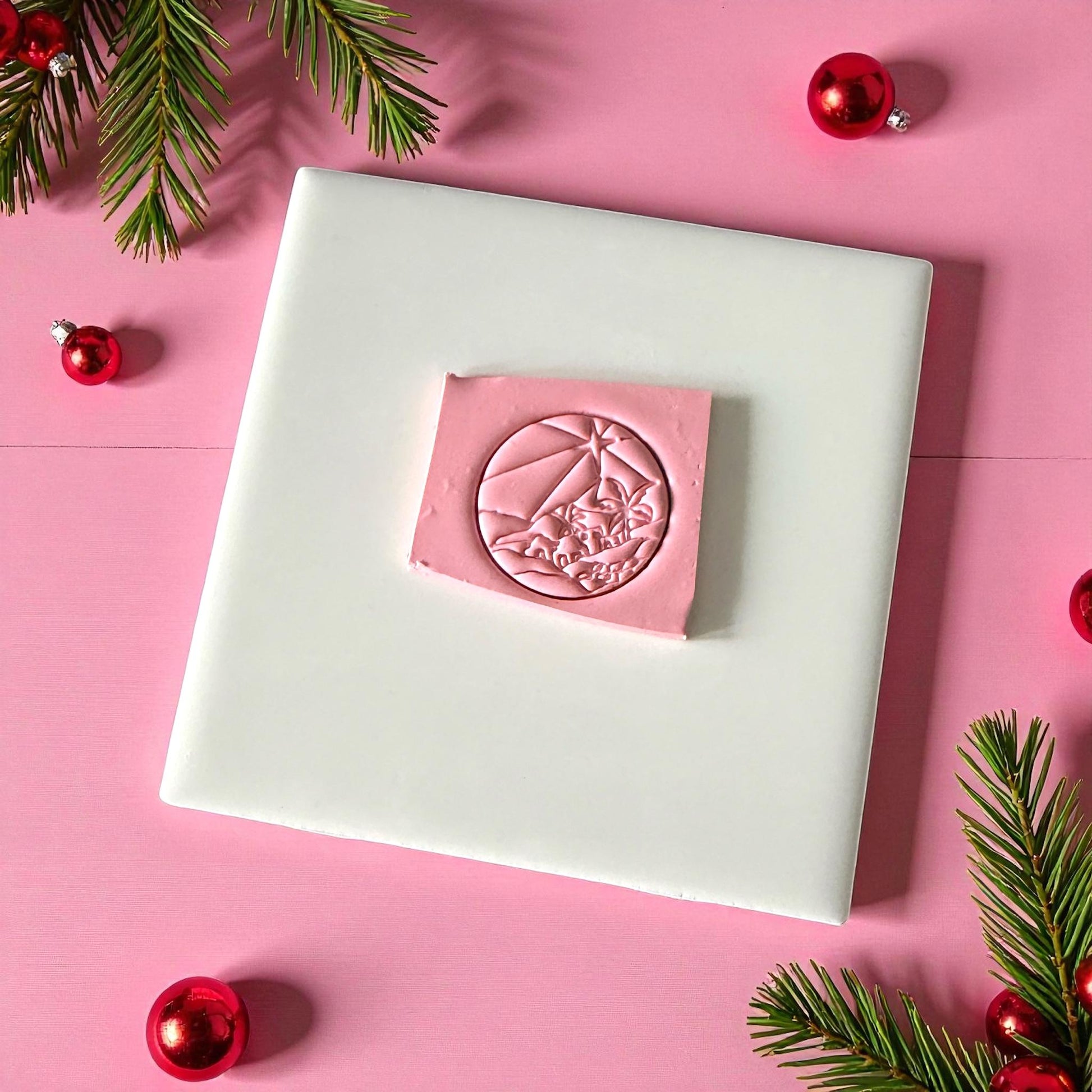 Pink clay imprint of Bethlehem town scene on white tile with pine sprigs and ornaments