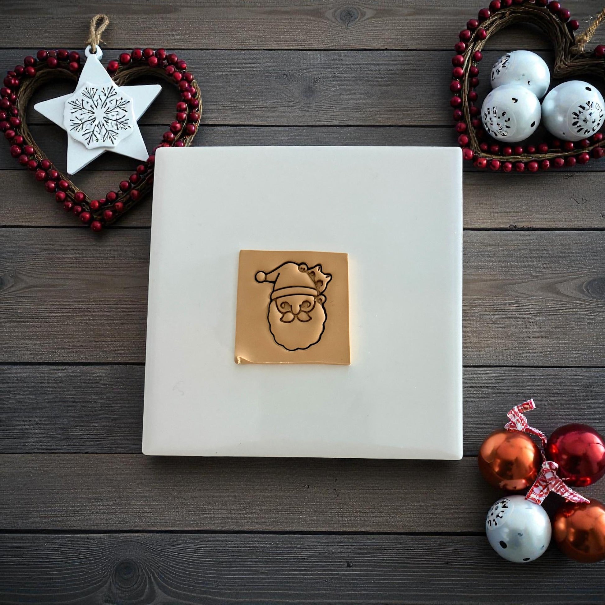 Santa face polymer clay imprint with cat peeking from hat on white tile with Christmas ornaments