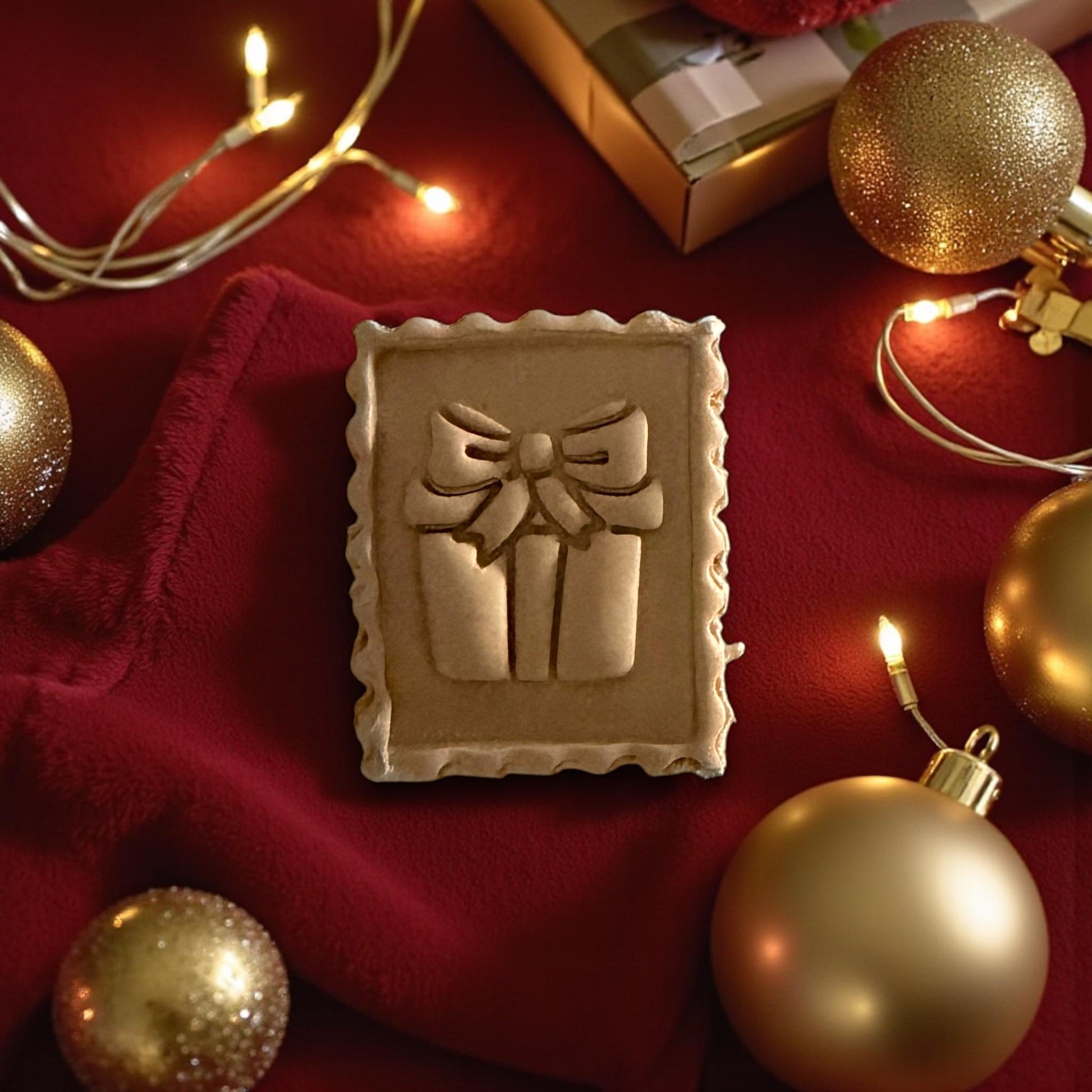Large gift stamp clay imprint on red fabric surrounded by Christmas ornaments and lights
