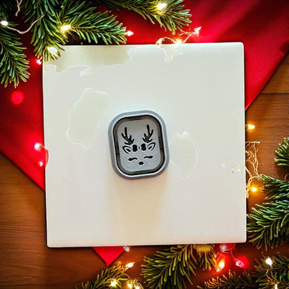 Small reindeer clay cutter centered on white tile with red fabric and holiday lights