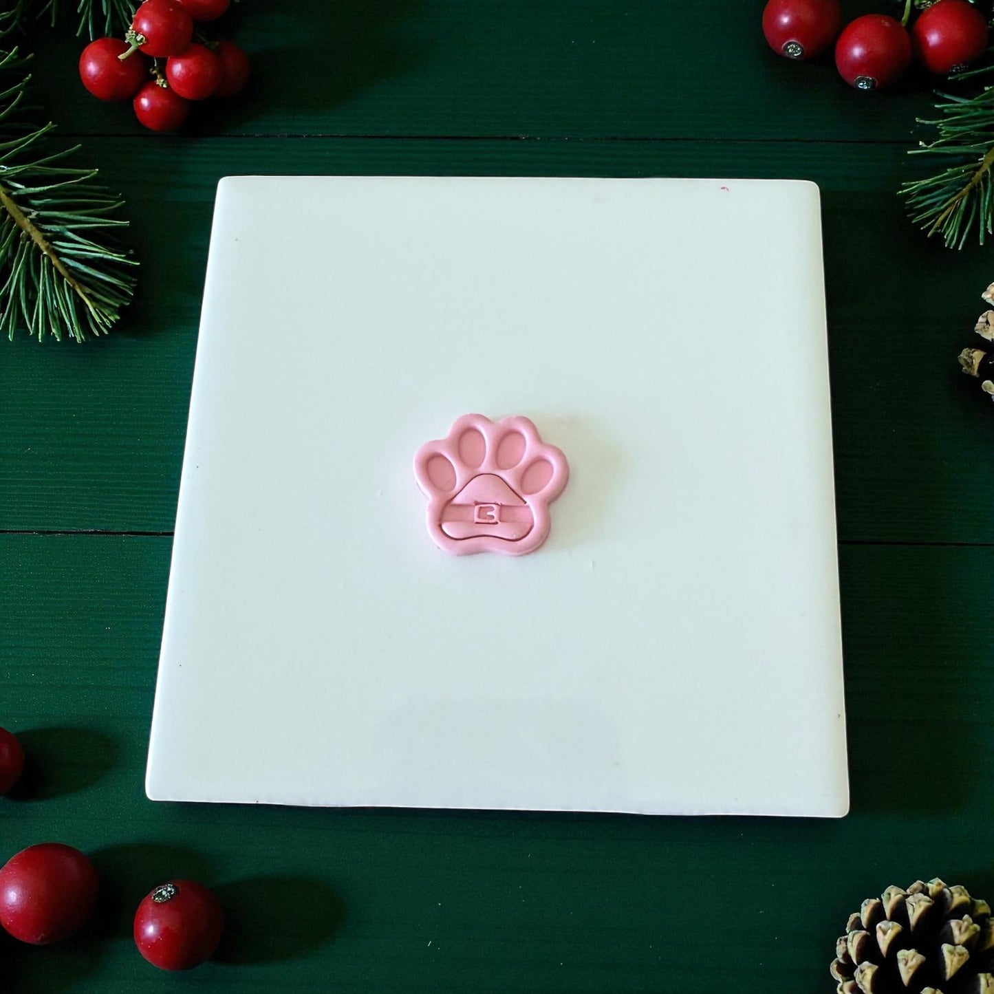 Pink Santa paw clay cutter centered on white tile with pine and berries