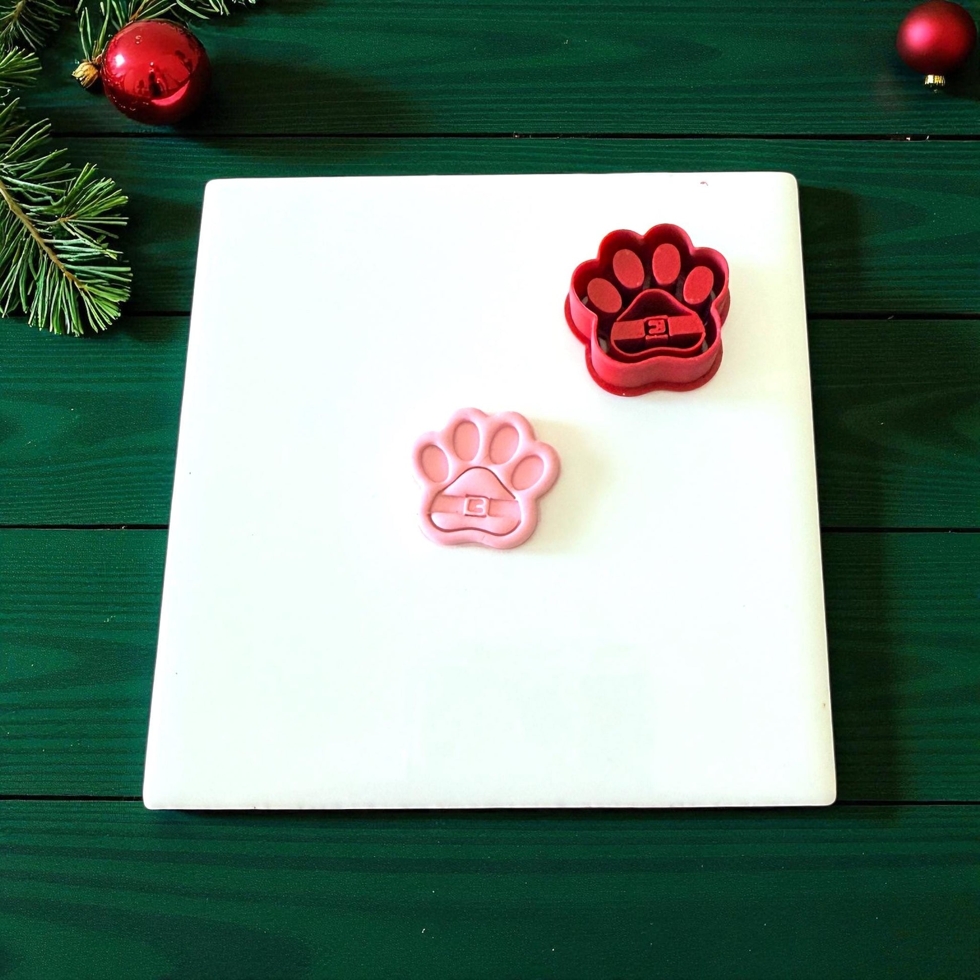 Red and pink Santa paw clay cutters displayed on white tile with pine accents