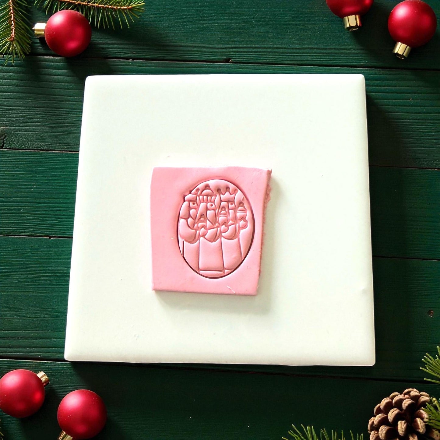 Pink Three Kings clay imprint on white tile surrounded by pine and red ornaments