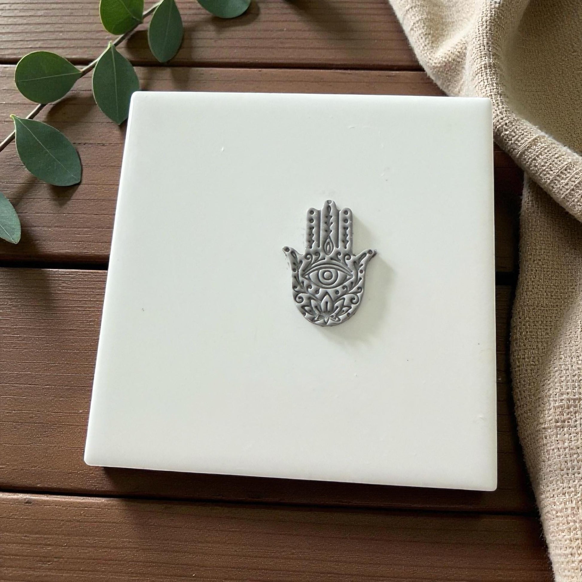 Gray Hamsa clay imprint centered on white tile with eucalyptus leaves