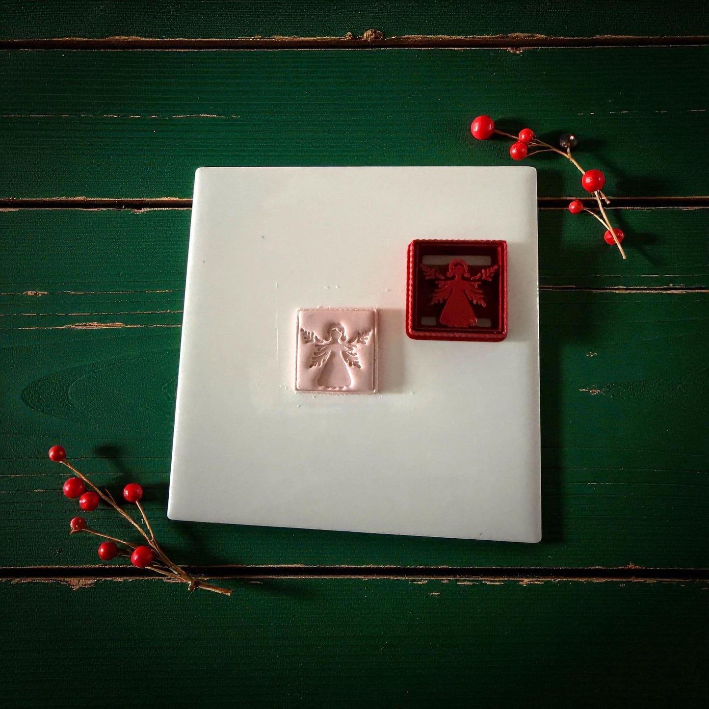 Pink angel clay imprint beside red square cutter on white tile with holiday decor