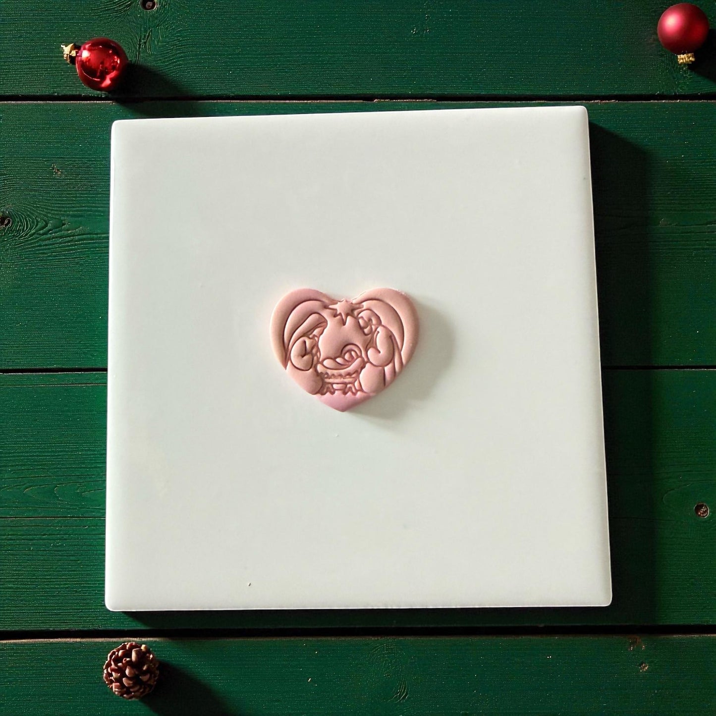 Pink heart nativity clay piece centered on white tile with green background and ornaments