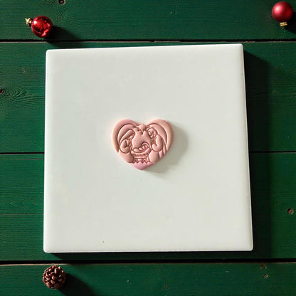 Pink heart nativity clay piece centered on white tile with green background and ornaments
