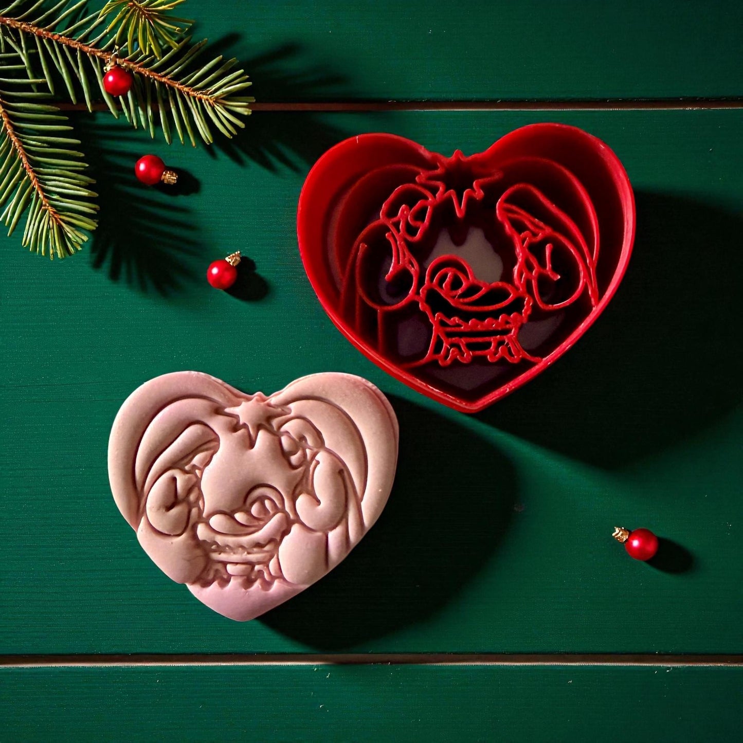 Red heart nativity cutter with pink clay imprint beside it on green wood with pine sprigs