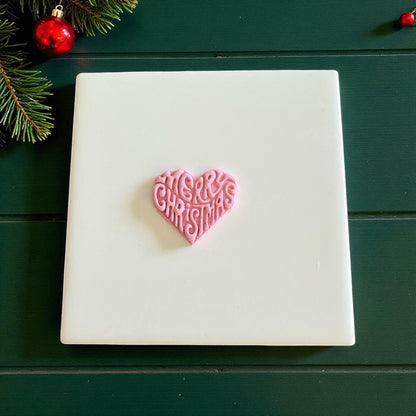 Pink heart clay piece with Merry Christmas text centered on white tile