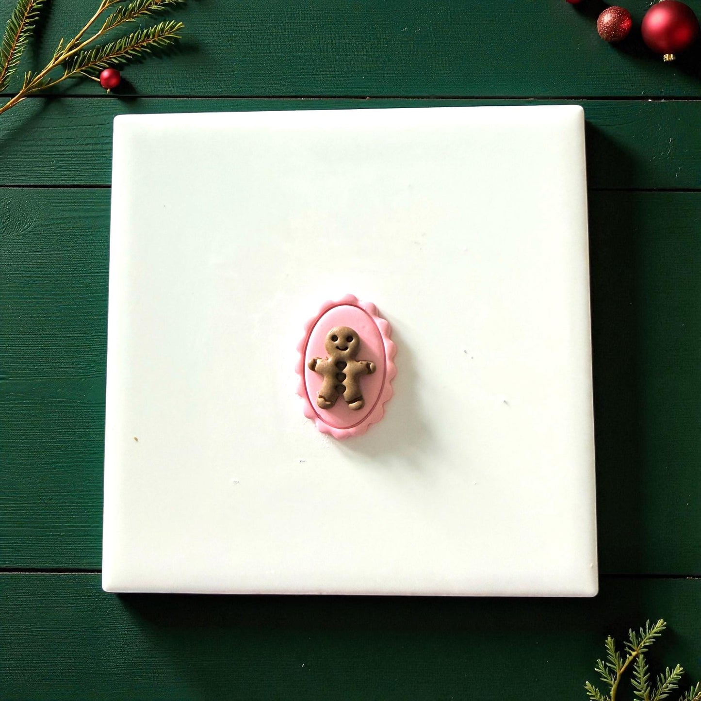 Gingerbread cameo clay piece centered on white tile surrounded by Christmas greenery