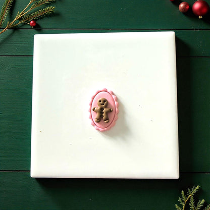 Gingerbread cameo clay piece centered on white tile surrounded by Christmas greenery