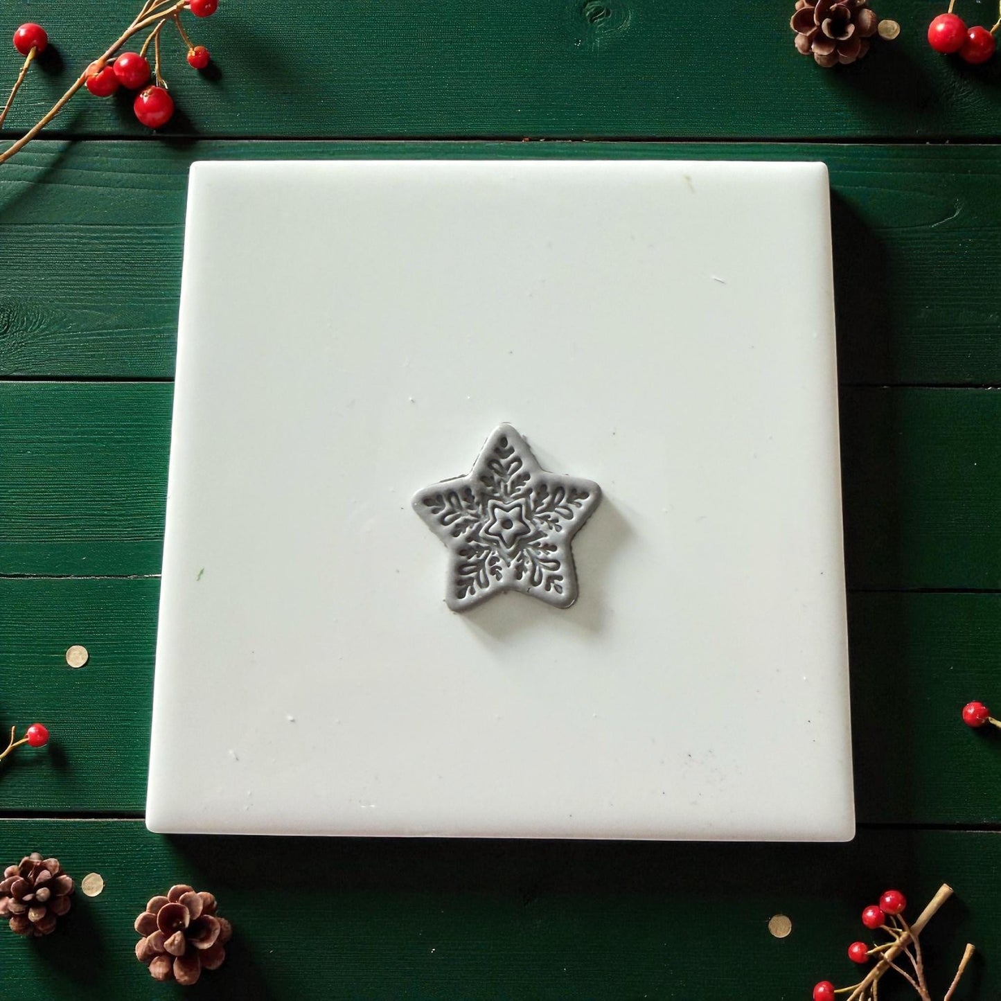 Single gray clay snowflake star on white tile with scattered pinecones and red berries