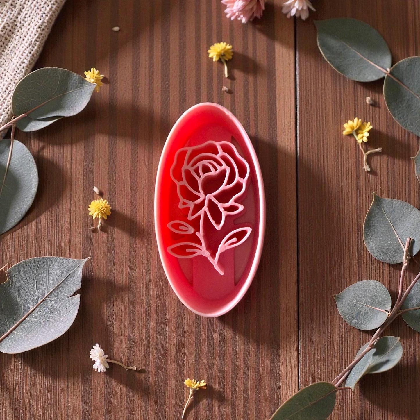 Pink oval rose polymer clay cutter on brown wood surrounded by eucalyptus and small yellow flowers