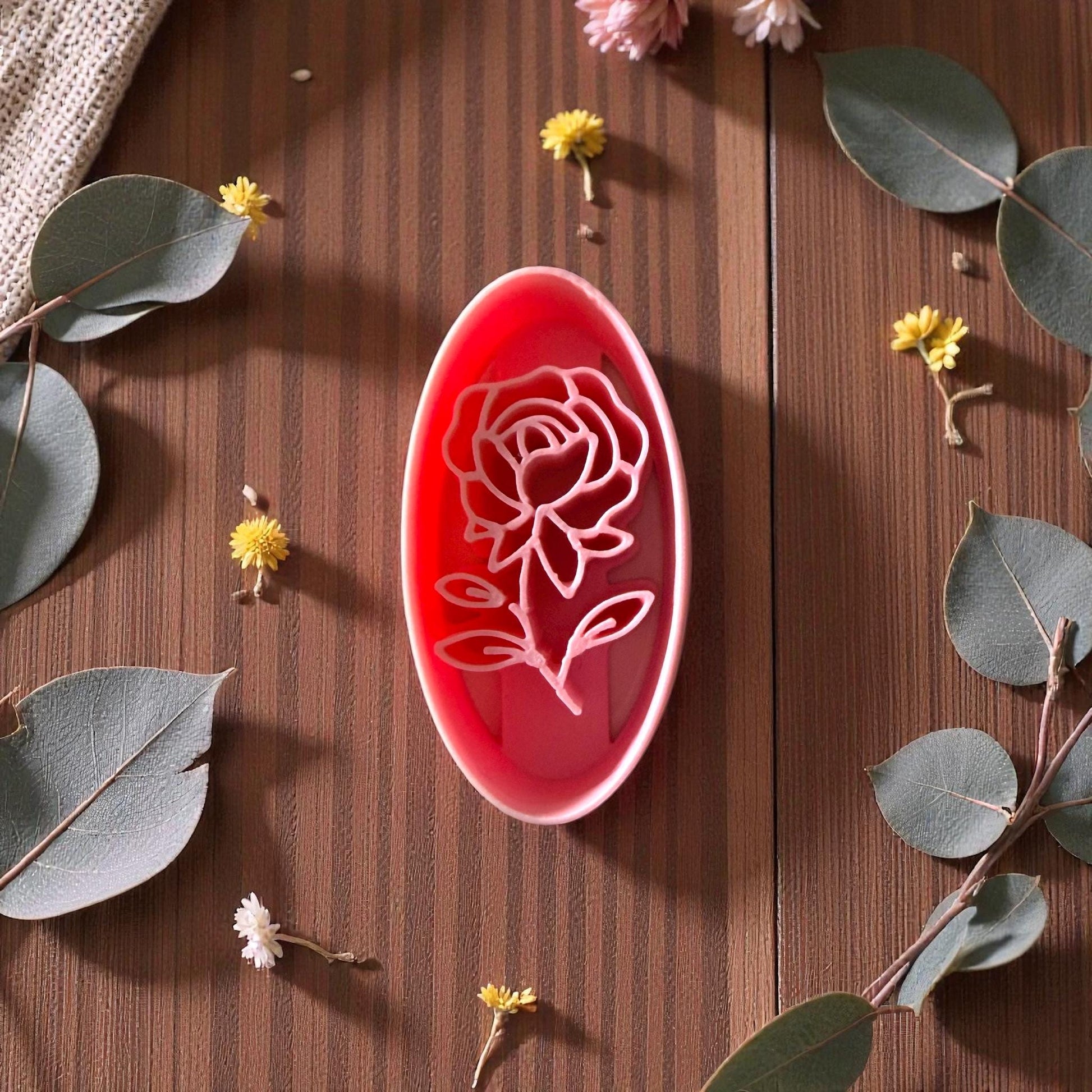 Pink oval rose polymer clay cutter on brown wood surrounded by eucalyptus and small yellow flowers