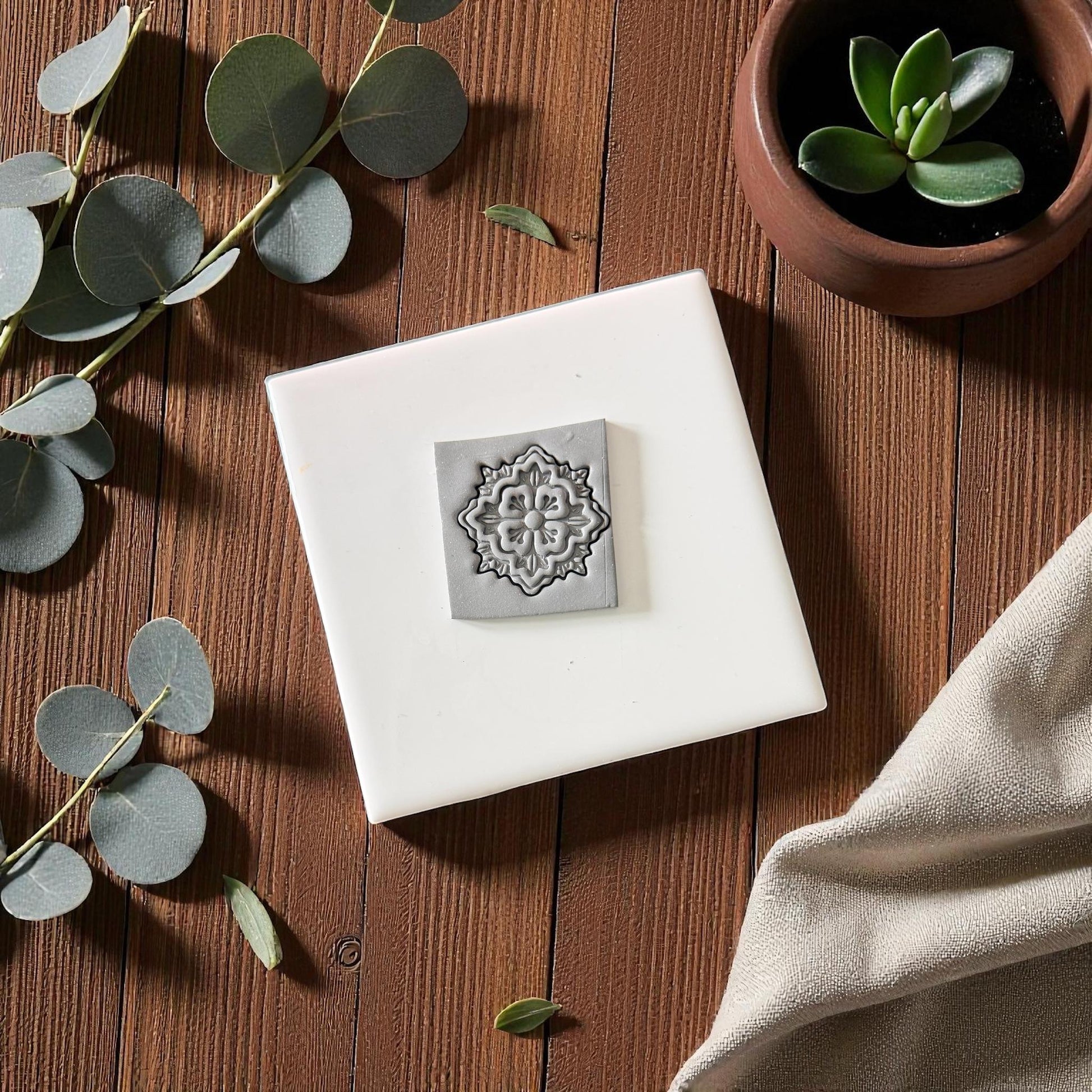 Gray floral clay imprint on square clay piece with eucalyptus and soft beige fabric