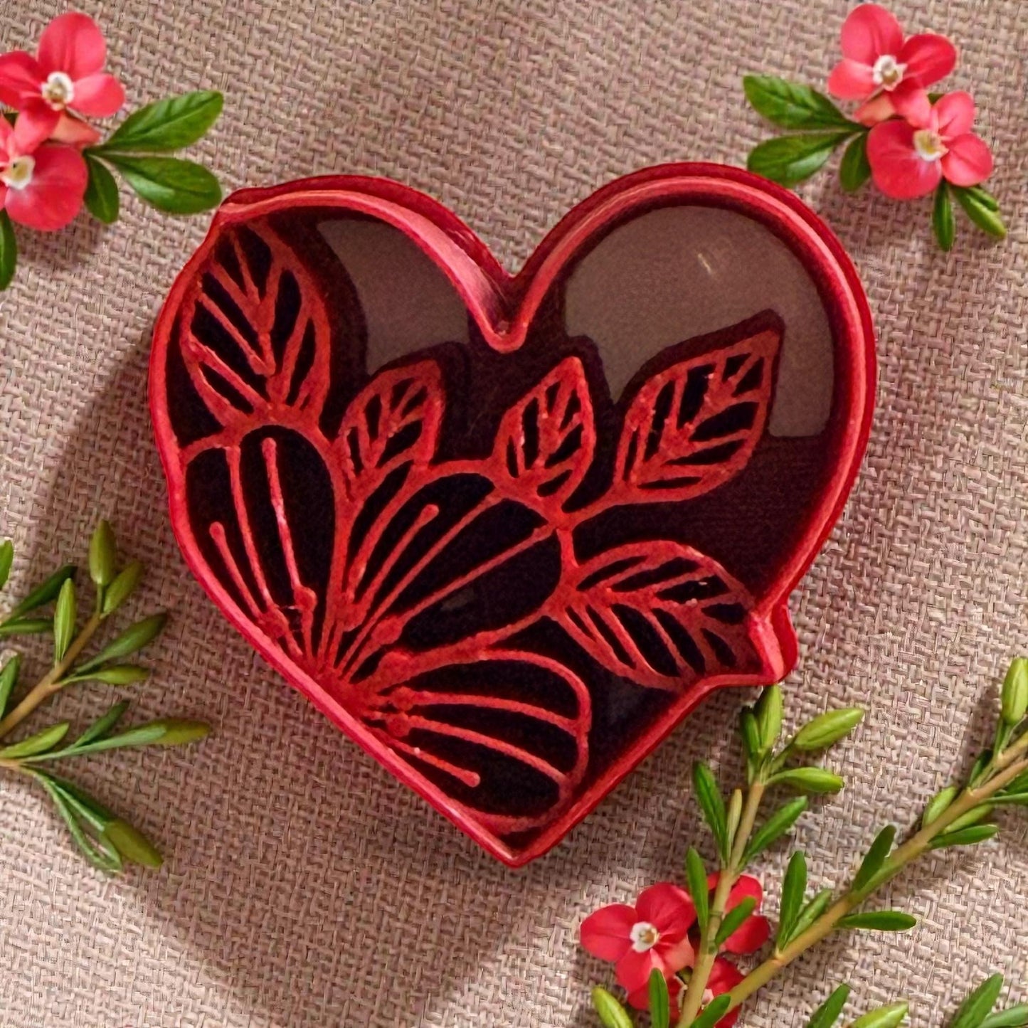 Red heart-shaped cutter with floral leaf design surrounded by pink flowers and greenery