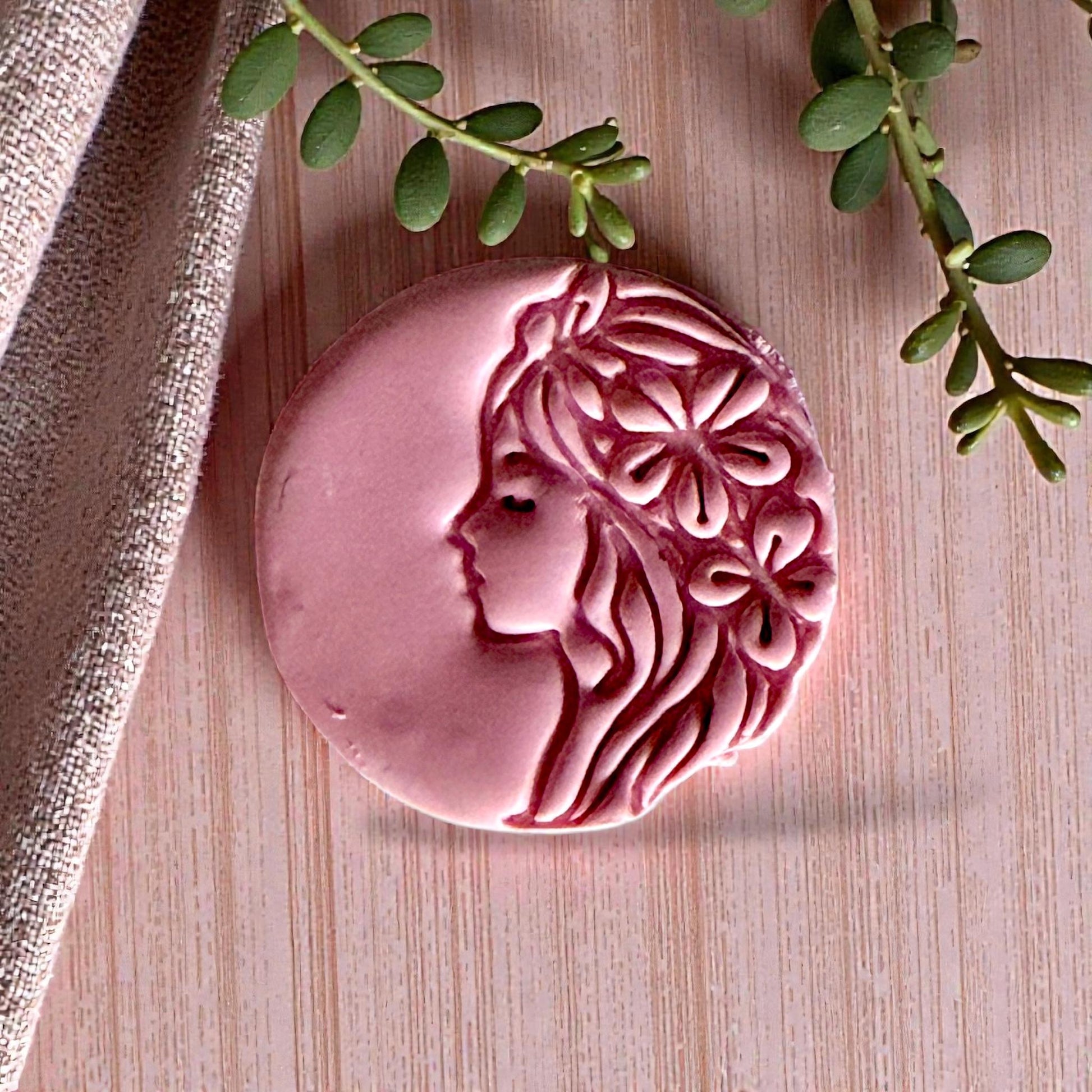 Pink clay impression of woman with flower hair beside textured linen