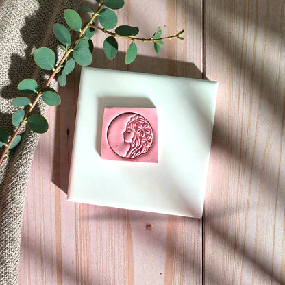 Pink woman profile clay impression with flowers on white tile surrounded by greenery