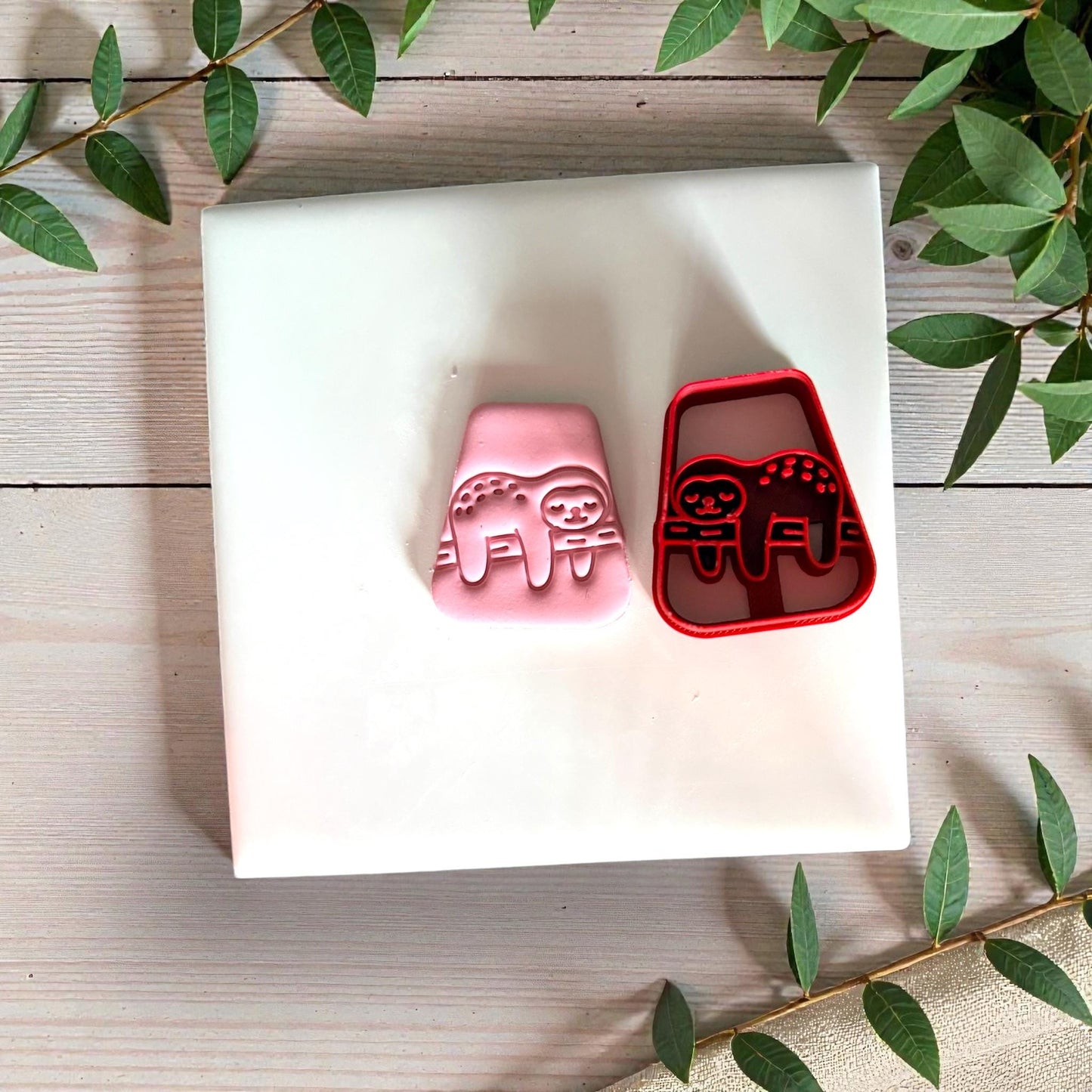 Red sloth cutter with pink clay impression beside it on white tile with greenery
