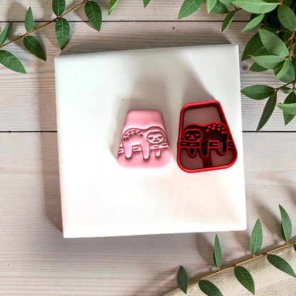 Red sloth cutter with pink clay impression beside it on white tile with greenery