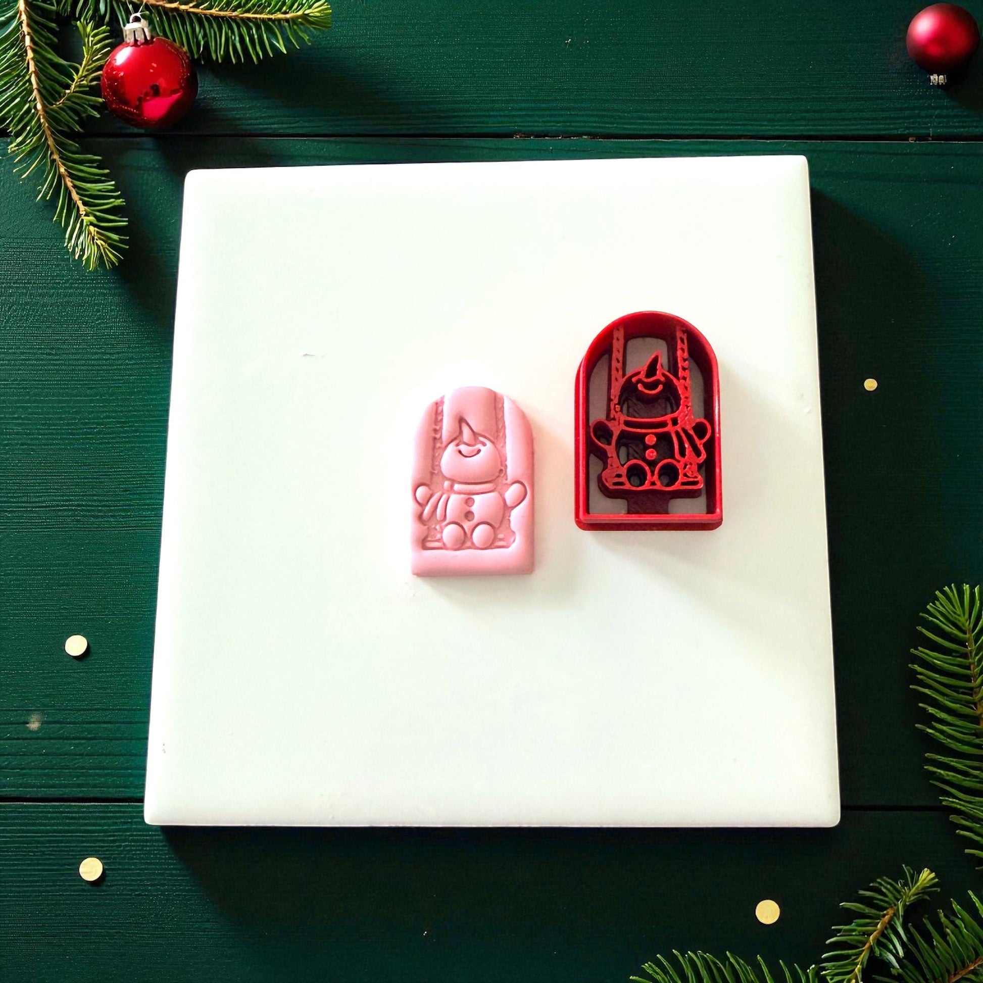 Pink clay imprint beside red snowman swing cutter on white tile
