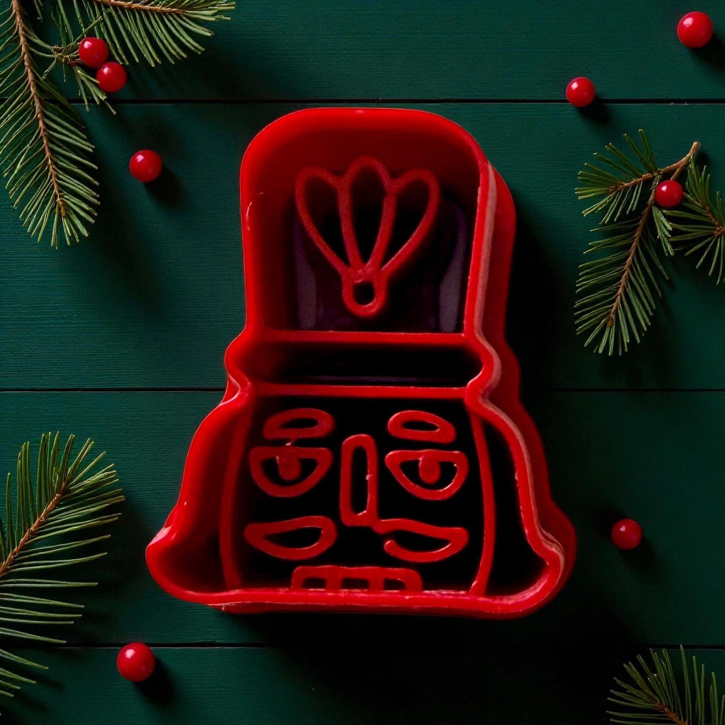 Red nutcracker head polymer clay cutter surrounded by pine branches and red ornaments