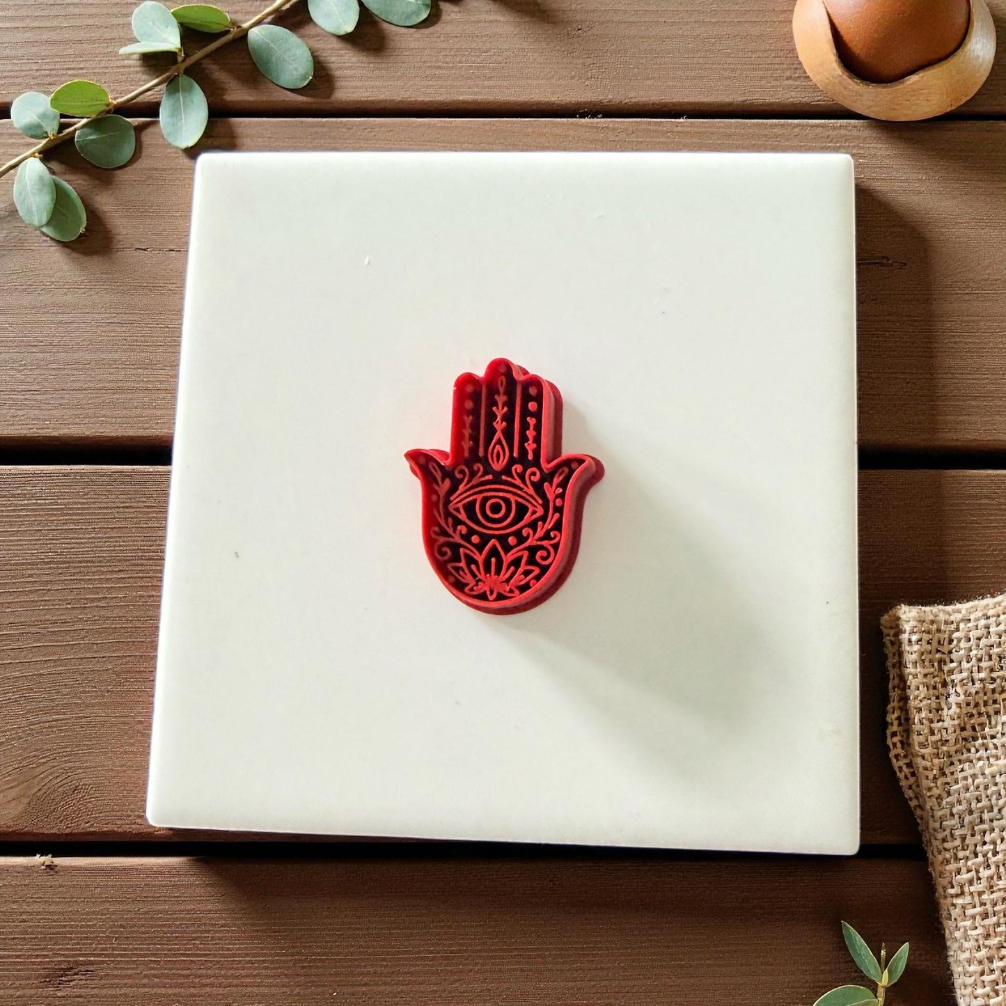 Red Hamsa Hand clay cutter centered on white tile with eucalyptus leaves