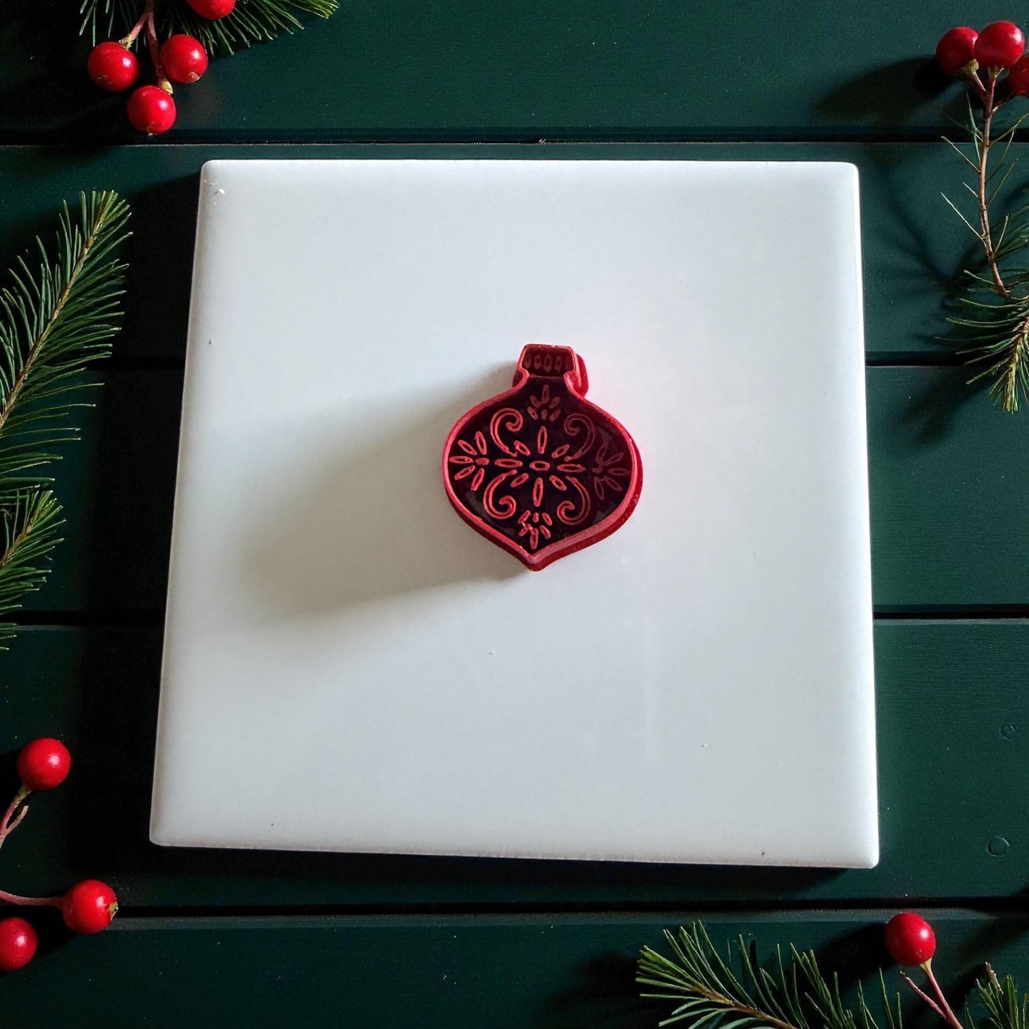 Red ornament cutter on white tile with greenery and berries