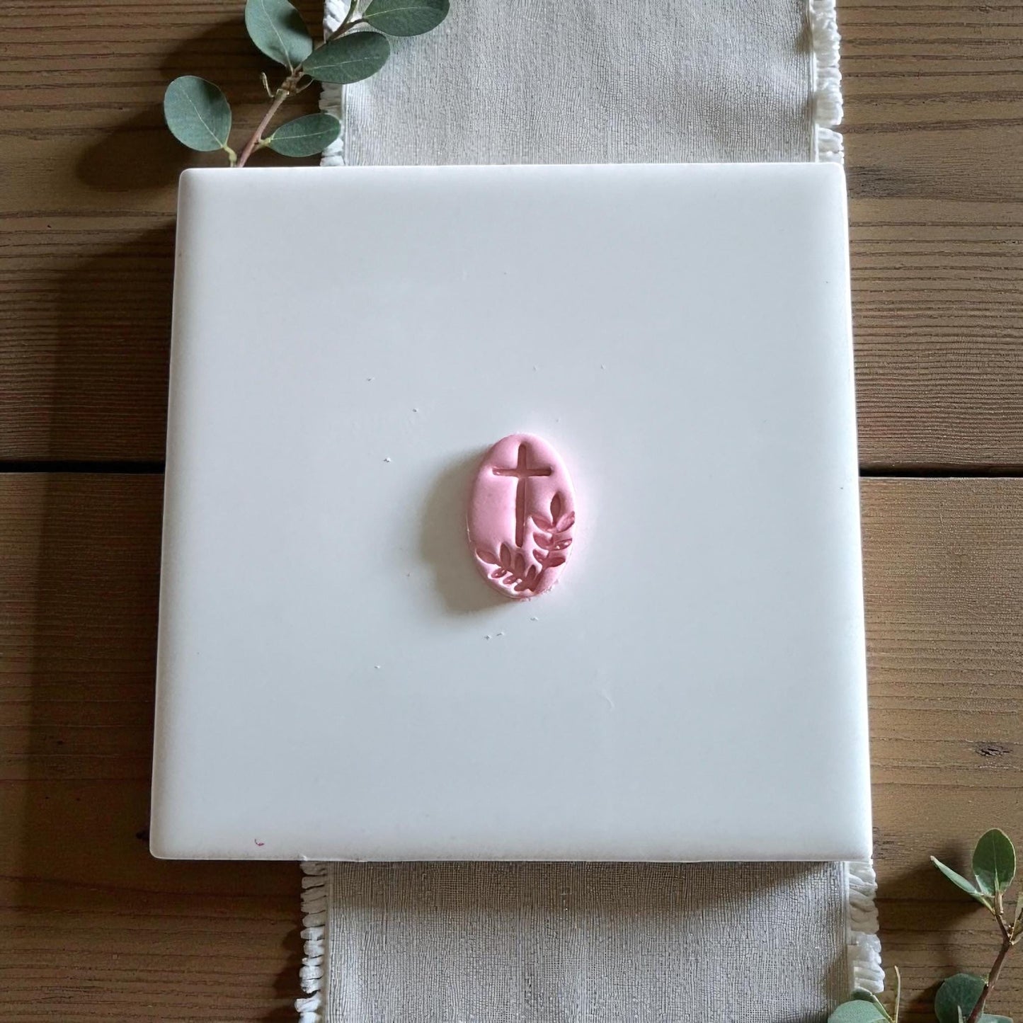 Small pink oval clay piece with cross and leaves detail on white tile
