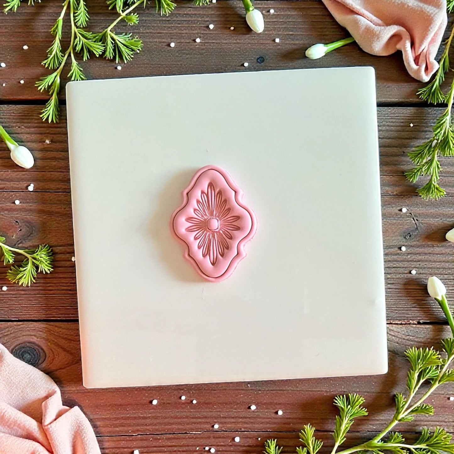 Pink ornate floral clay design centered on white tile with fabric and greenery
