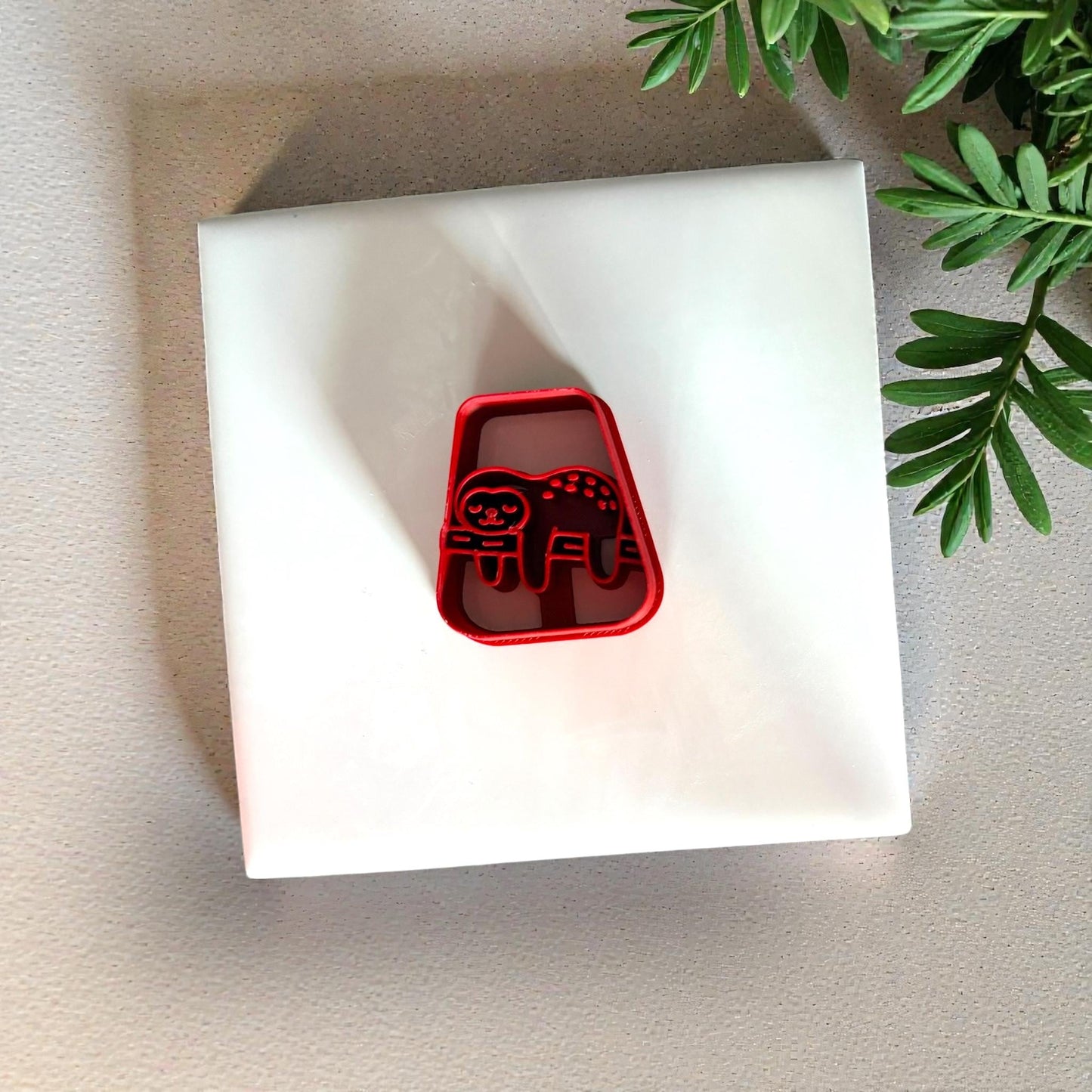 Red sloth clay cutter centered on white tile with green plant above