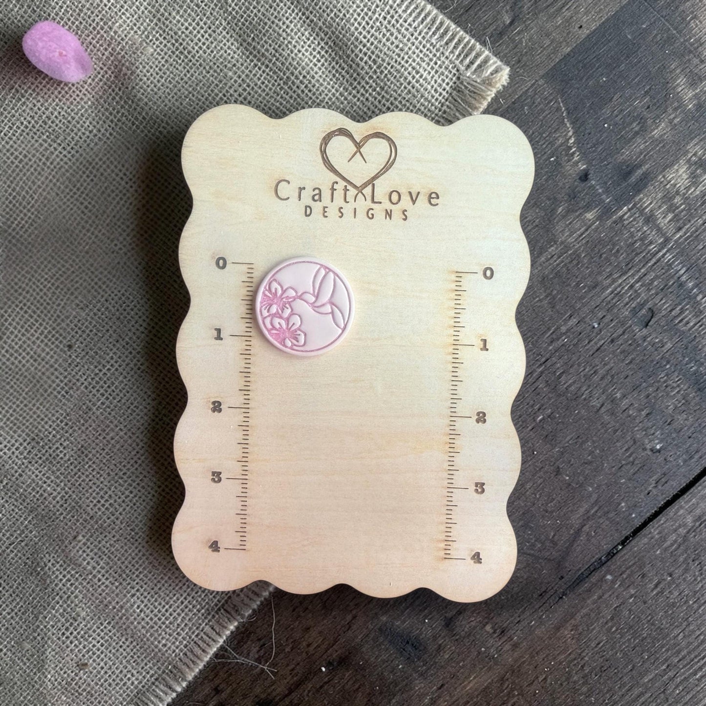 Finished round clay piece featuring a hummingbird drinking from a flower, displayed on a wooden measuring board.
