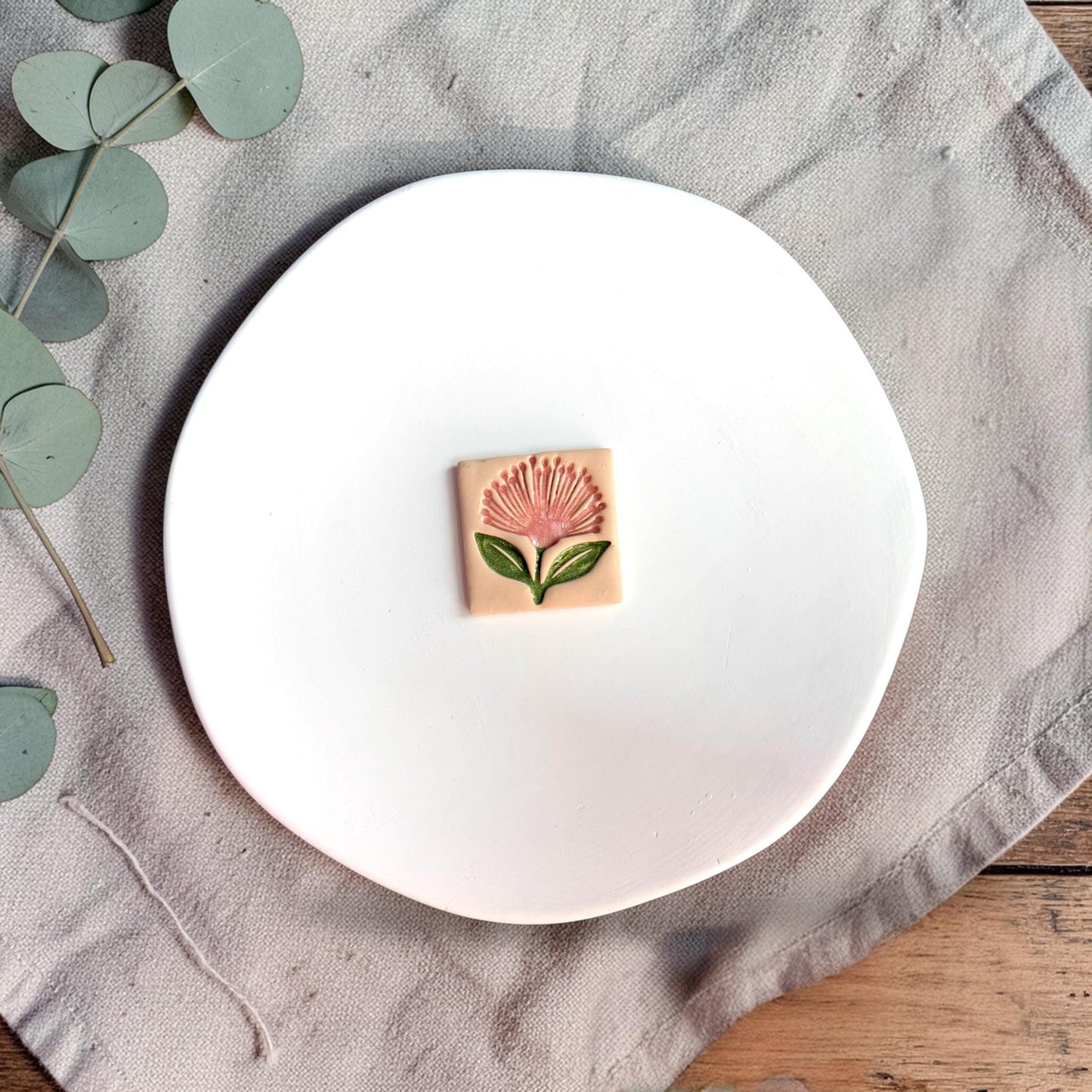 Finished clay piece showing a pink pohutukawa flower with green leaves on a square background placed on a white plate.