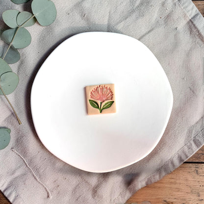 Finished clay piece showing a pink pohutukawa flower with green leaves on a square background placed on a white plate.