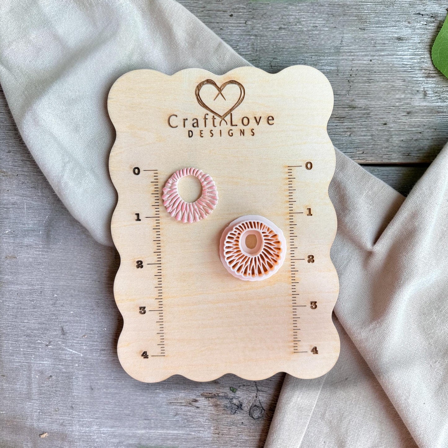Cutter and finished clay pieces displayed on a wooden measuring board with ruler markings and soft neutral fabric.
