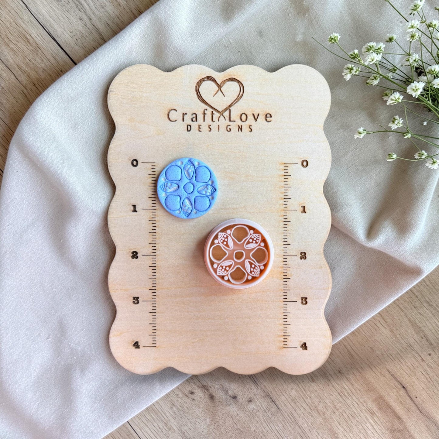 Two round clay pieces with sea shell floral pattern in blue and terracotta displayed on a wooden measuring board with ruler markings.