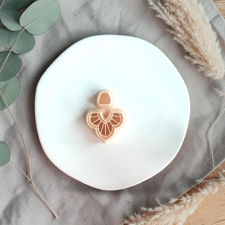 Clay cutter with scalloped fan wing design and small top piece placed on a white plate with neutral fabric and dried grass.