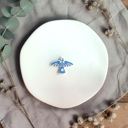 Small blue clay piece with intricate bird pattern placed on a white plate with neutral fabric and dried florals.