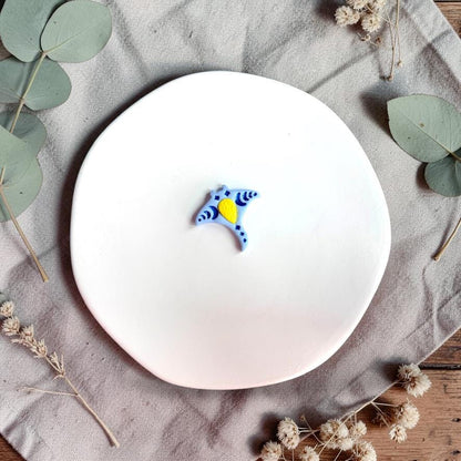 Small blue manta ray shaped clay piece with decorative pattern placed on a white plate with neutral fabric and dried flowers.