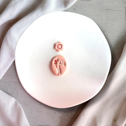 Feminine flamingo polymer clay earring design with small flower topper displayed on white plate with soft fabric background.