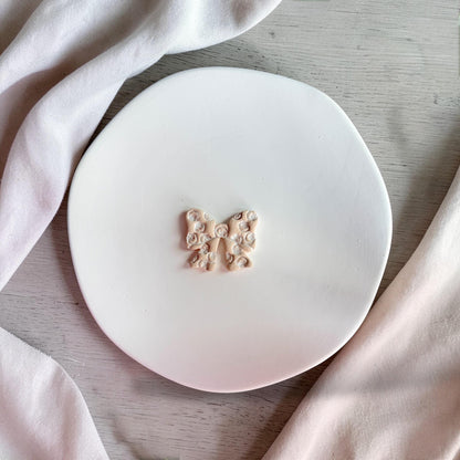 Finished seashell bow polymer clay earring piece on white plate with soft neutral fabric background.