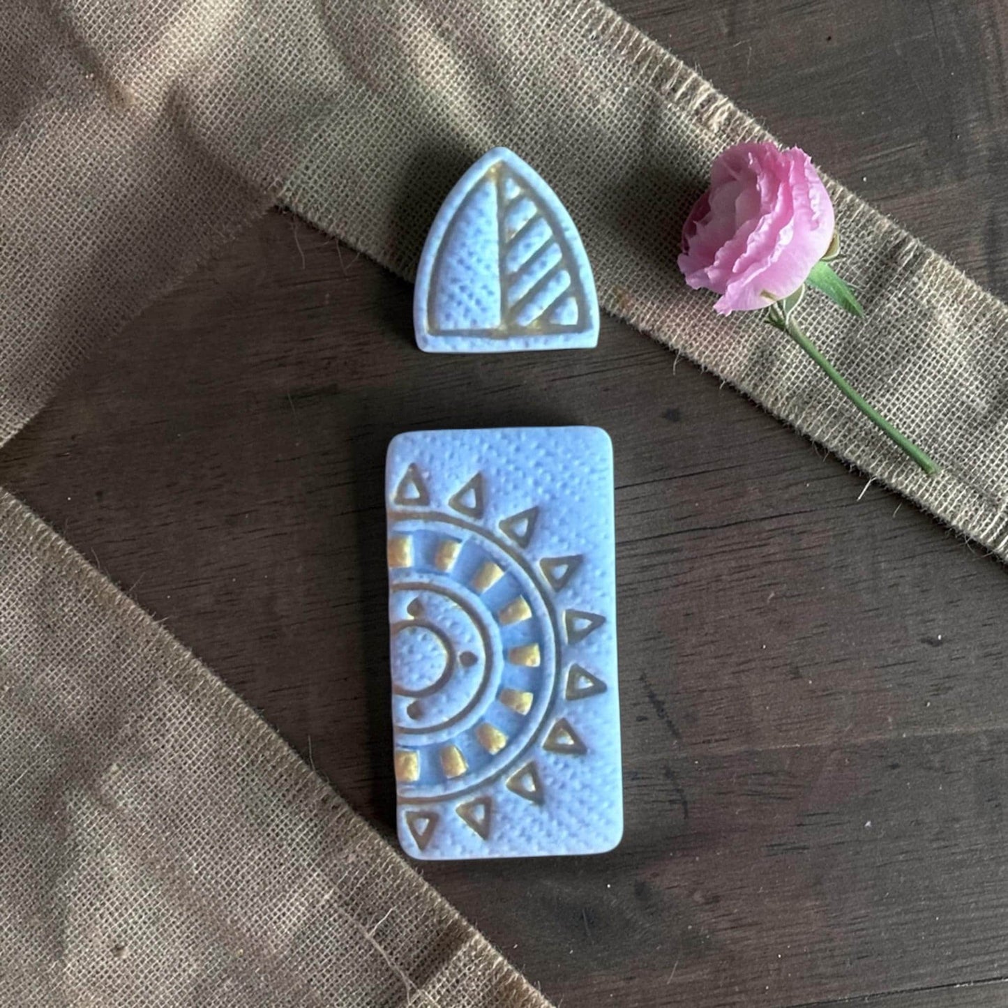 Finished blue clay earrings with triangular stud and textured sunburst dangle on linen fabric