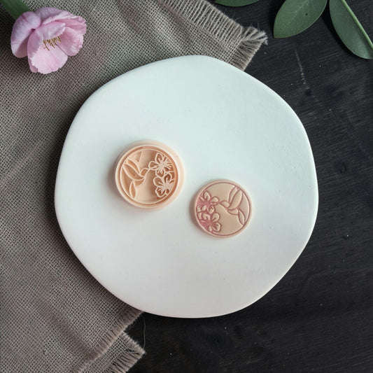 Two round clay pieces with hummingbird and flower design displayed on a white circular board with soft styling.