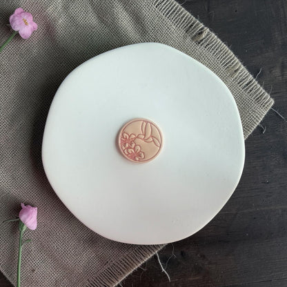Single round polymer clay hummingbird placed on a white display board with burlap fabric and floral accents