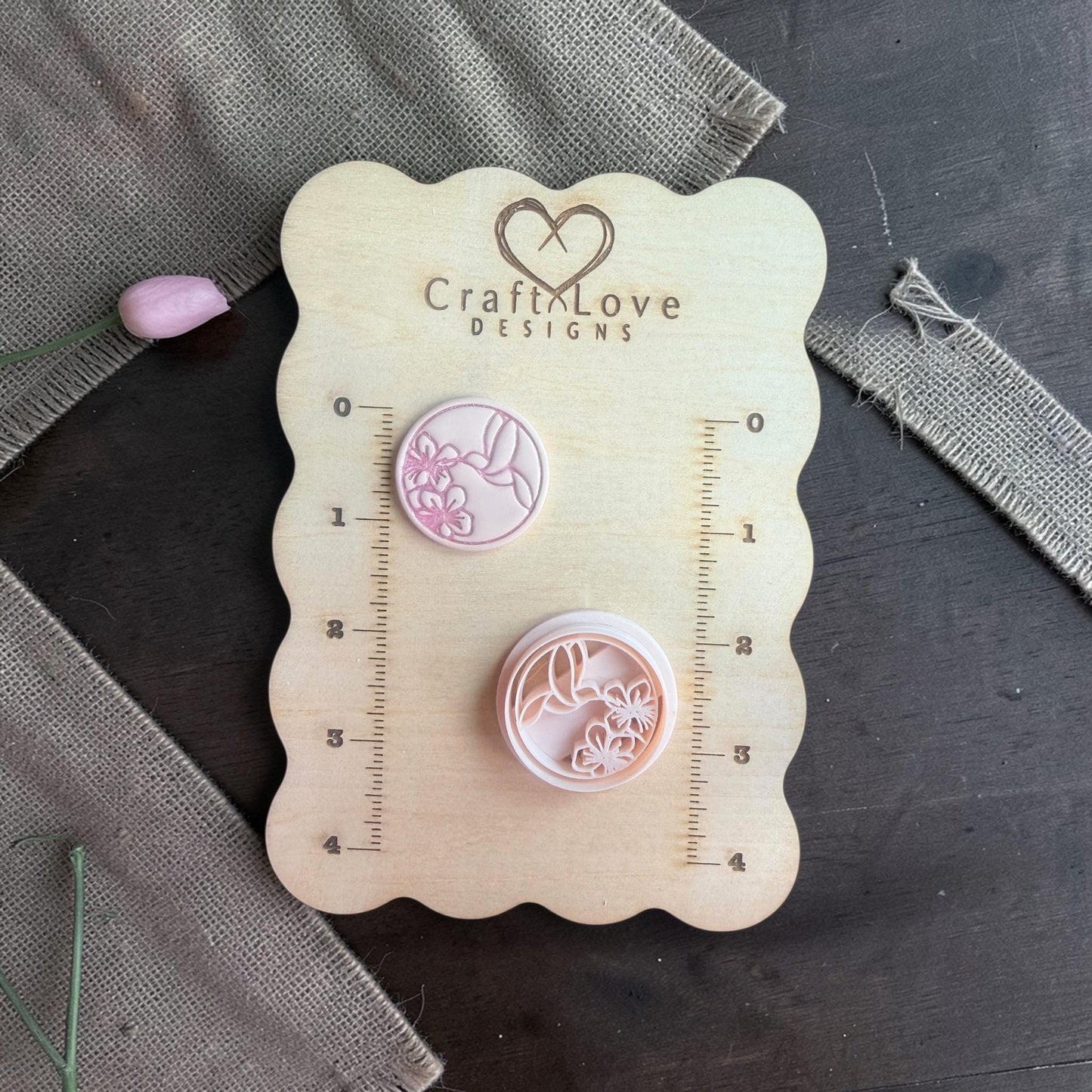 Round polymer clay cutter with hummingbird and flower design shown with matching clay piece on a wooden measuring board with burlap fabric.