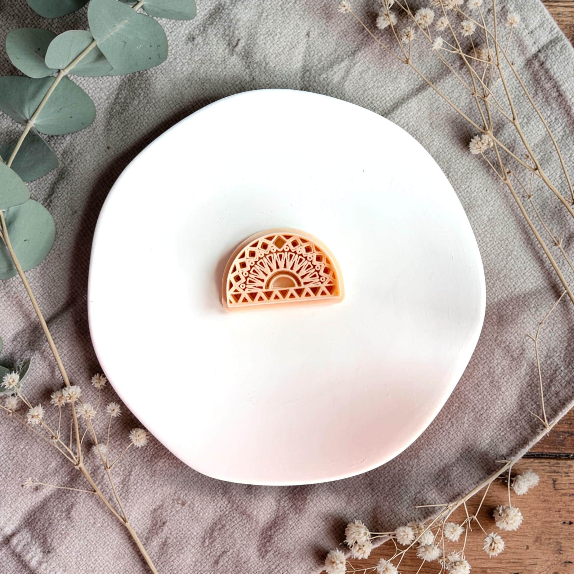 Small arch-shaped clay piece with intricate geometric sunburst pattern displayed on a white round plate with neutral fabric and greenery.